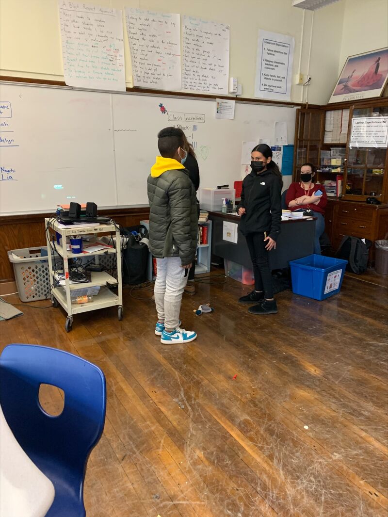 The image shows a classroom setting. Two students are standing in front of a whiteboard, one wearing a green jacket and the other a black hoodie. A third person is seated at a desk in the background. There are shelves, a blue bin, and a cart with items on it. The room appears to be well-lit.